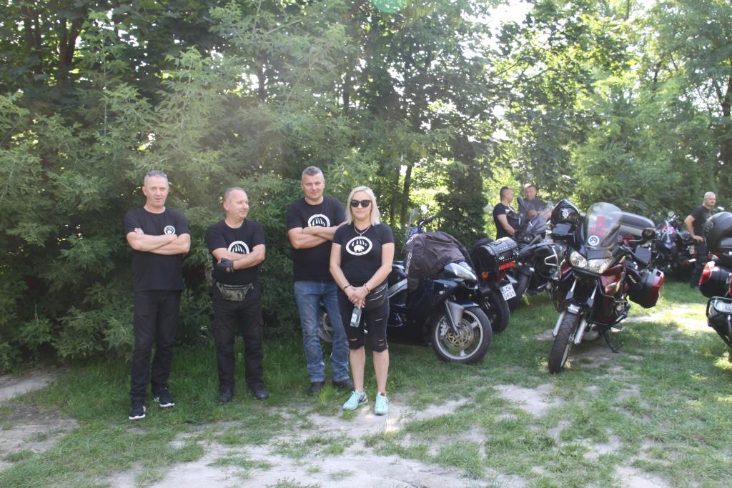 Motocykle ustawione na trawie, obok nich właściciele. Na pierwszym planie 4 osoby w czarnych koszulkach.